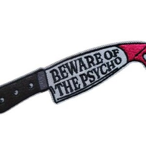 May include: A white embroidered patch shaped like a knife with a black handle and a red dripping blood effect. The text on the patch reads "Beware of the Psycho".