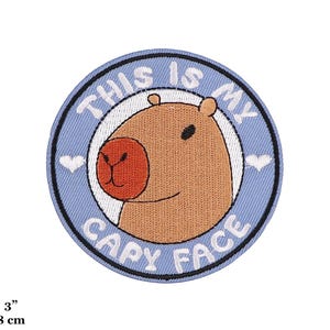Peut inclure: Écusson brodé avec un visage de capybara de dessin animé. Le patch rond a une bordure bleu clair et le texte "THIS IS MY CAPY FACE". Le capybara est marron avec un nez rouge. Le patch mesure 8 cm de diamètre.