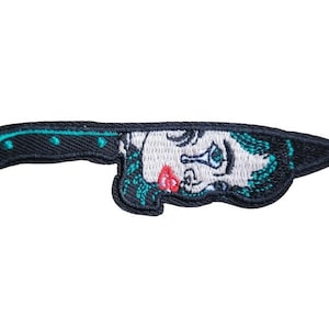May include: An embroidered iron-on patch of a black knife with a woman's face peeking out from the blade. The woman has blue eyes, red lips, and green hair. The patch is 3.5 inches by 1 inch.