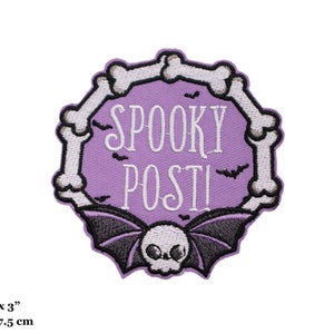 Könnte beinhalten: Ein lila gestickter Bügel-Patch mit einem Totenkopf- und Fledermaus-Design. Der Patch ist von einem Kreis aus weißen Knochen umgeben und trägt den Schriftzug "Spooky Post!" in weißer Schrift. Der Patch ist 7,5 cm x 7,5 cm groß.