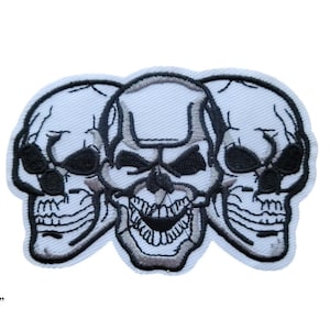 May include: Embroidered patch featuring three white skulls with black details. The skulls are arranged in a row, with the middle skull facing forward and the two outer skulls facing slightly outward. The patch measures 9 cm by 6 cm.