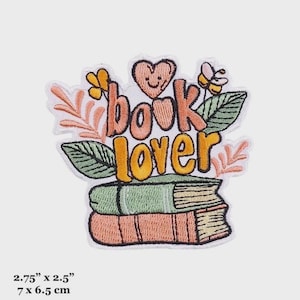 Puede incluir: Parche bordado para planchar con una pila de libros, un corazón con una carita sonriente y el texto "book lover" en naranja, rosa y verde. El parche mide 7 cm por 6,5 cm.