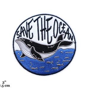 Könnte beinhalten: Ein runder, weißer gestickter Aufnäher mit blauem Hintergrund. Der Aufnäher zeigt einen Wal, der im Meer schwimmt, mit dem Text "Save The Ocean" in schwarzen Buchstaben.