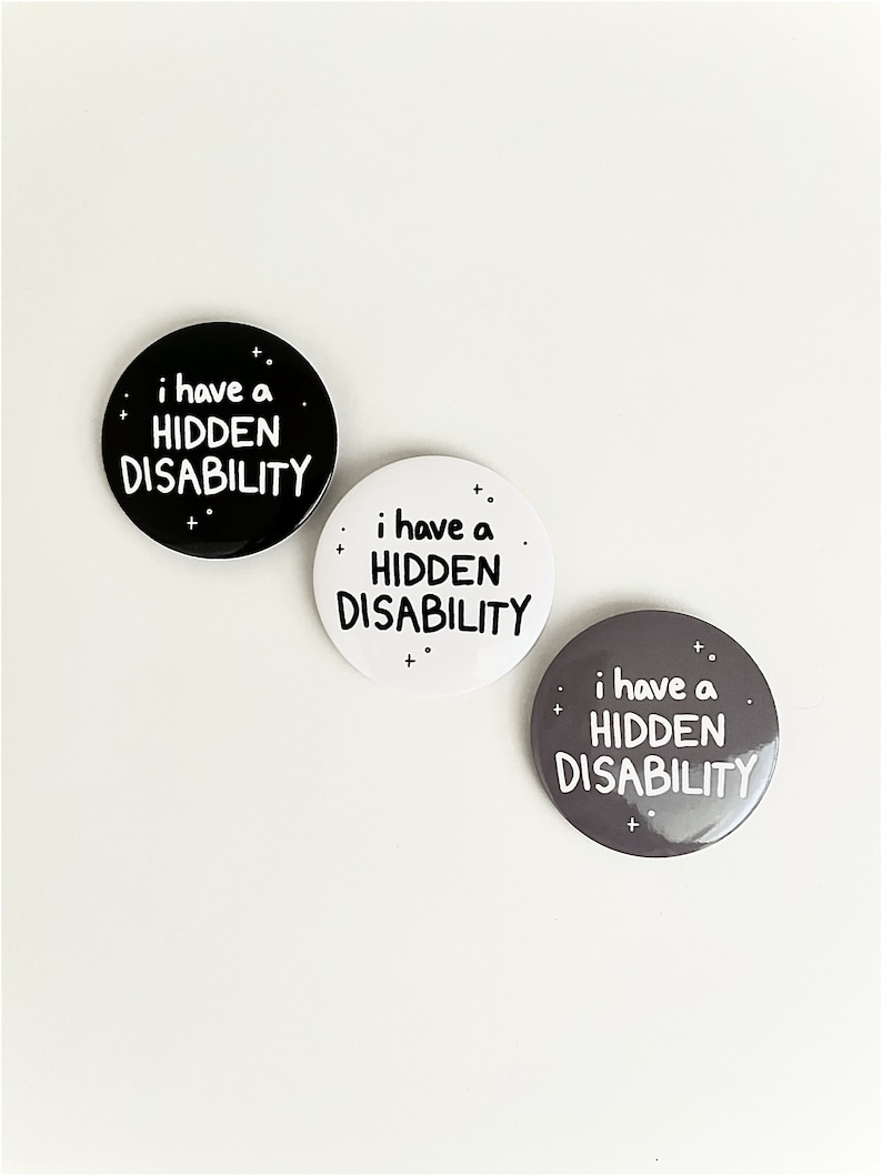 Puede incluir: Tres botones redondos con el texto "i have a HIDDEN DISABILITY" en letras blancas sobre fondo negro, blanco y gris.