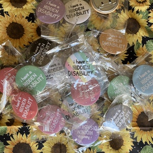 Puede incluir: Una colecci&oacute;n de botones coloridos con el texto "Tengo una discapacidad invisible" en varios colores y fuentes. Los botones est&aacute;n empaquetados en bolsas de pl&aacute;stico transparentes y se muestran sobre un fondo de tela con girasoles.