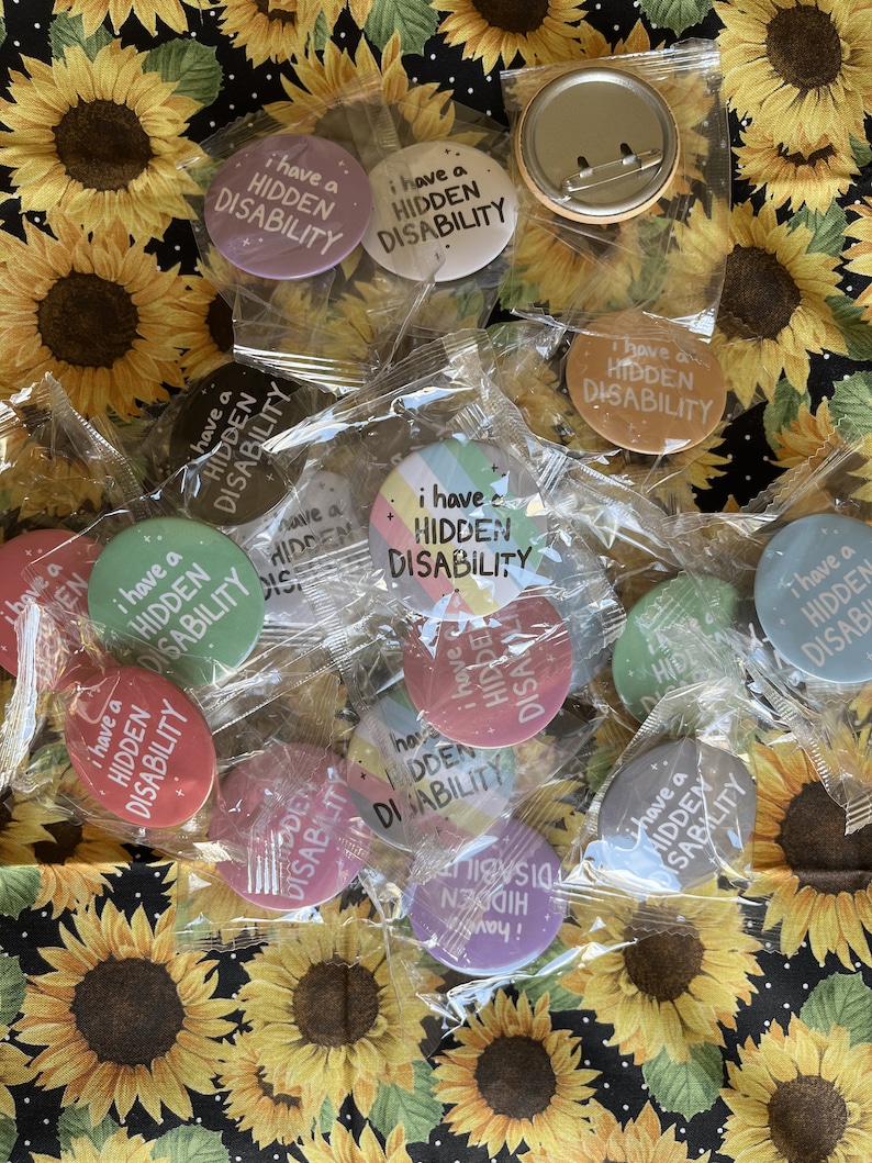 Puede incluir: Una colecci&oacute;n de botones coloridos con el texto "Tengo una discapacidad invisible" en varios colores y fuentes. Los botones est&aacute;n empaquetados en bolsas de pl&aacute;stico transparentes y se muestran sobre un fondo de tela con girasoles.