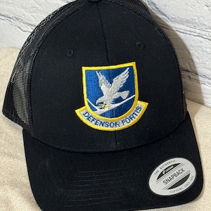 May include: Black and yellow trucker hat with a blue and white embroidered patch that says "DEFENSOR FORTIS". The hat has a mesh back and a black snap closure. The hat is labeled "THE AUTHENTIC SNAPBACK YUPOONG SINCE 1974".