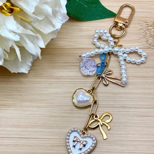 May include: A gold-toned keychain with a pearl-beaded bow, a clear rose charm, and a chain of charms. The charms include a heart-shaped pendant with a pearl center and a bow, and a gold bow charm. The keychain is on a wooden surface.