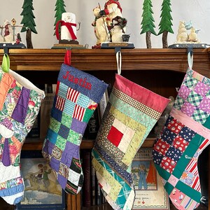 May include: Four colorful patchwork Christmas stockings hang on a wooden mantel. The stockings are decorated with various fabrics and patterns, including plaid, floral, and geometric designs. The stockings are personalized with the names "Justin", "Lucy", and two other names that are not visible in the image.