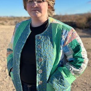 May include: A person wearing a green and blue quilted jacket with a paisley pattern. The jacket has a button closure and pockets. The person is standing in a desert landscape.