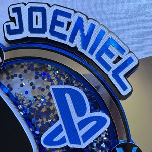 May include: A blue and white PlayStation themed cake topper with the name "Joeniel" in blue block letters. The cake topper features a PlayStation logo, a controller, and a blue and silver glitter background.