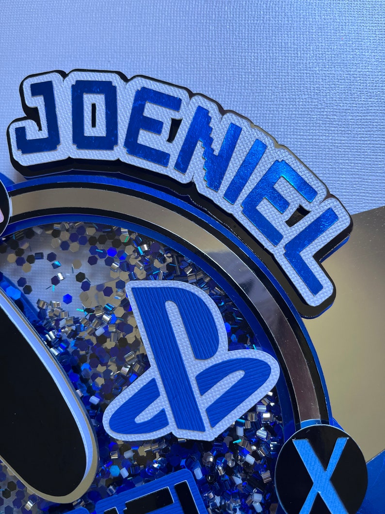 May include: A blue and white PlayStation themed cake topper with the name "Joeniel" in blue block letters. The cake topper features a PlayStation logo, a controller, and a blue and silver glitter background.
