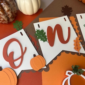 Puede incluir: Una pancarta festiva que deletrea "ONE" con letras naranjas, hojas verdes y acentos de calabaza. La pancarta está colocada sobre un fondo de papel naranja y marrón, con calabazas decorativas en el fondo.