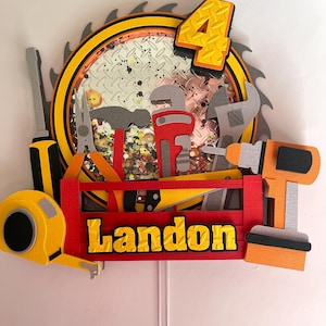 Könnte beinhalten: Ein farbenfroher Tortenaufsatz zum Geburtstag mit einem Sägeblatt, Werkzeugen und der Zahl 4. Der Name "Landon" ist in gelben Buchstaben auf einer roten Werkzeugkiste dargestellt. Das Sägeblatt hat einen gelb-schwarzen Rand.