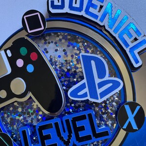 May include: A blue and silver cake topper with a video game controller, a PlayStation logo, and the text "Joeniel Level 10".