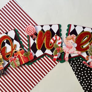 May include: A festive banner with the word "ONE" in red glitter letters, featuring a nutcracker, gifts, a ballerina, and a Christmas tree. The banner has a black and white checkered pattern and is trimmed with green scalloped edges.