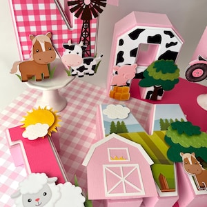 Puede incluir: Decoración de cumpleaños con temática de granja en rosa y blanco, con la letra "N" y el número "1". Las decoraciones incluyen un molino de viento, un granero, una vaca, un caballo, una oveja y un tractor. El fondo es un patrón de cuadros rosas.