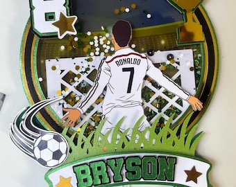 May include: A layered paper cake topper featuring a soccer theme with a central figure of a player in a white jersey with Ronaldo 7, a white soccer goal, a gold trophy, a soccer ball, the number 8, and a banner with the name BRYSON in green.