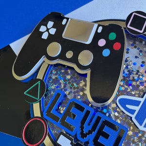 May include: A blue and black cake topper with a black video game controller, a blue and black "LEVEL 10" sign, and silver glitter.