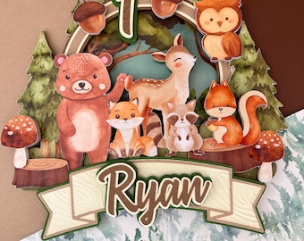 Può includere: Decorazione per torta a tema bosco con orso, cervo, volpe, procione, scoiattolo e gufo. Il numero "1" e il nome "Ryan" sono visualizzati. Il design include alberi, ghiande e funghi, con uno sfondo di foresta.