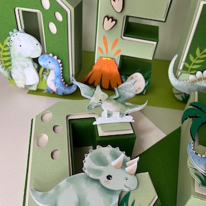 Könnte beinhalten: Eine dreidimensionale grün-weiße Dekoration zum Thema Dinosaurier, die das Wort "ONE" buchstabiert. Die Buchstaben sind mit Cartoon-Dinosauriern, einem Vulkan und Blättern verziert. Die Farbpalette umfasst Grün-, Weiß-, Orange- und Blautöne.