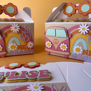 May include: Three pastel-coloured party favour boxes with a groovy 70s theme. Each box features a rainbow, peace sign, and a VW van with a peace sign. The boxes are decorated with flowers and sunglasses. One box has the name "Elise" on the front.