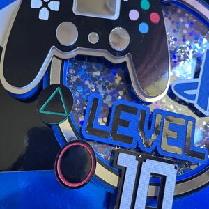 May include: A blue and silver cake topper with a black video game controller, a triangle, a circle, and the text "LEVEL 10".