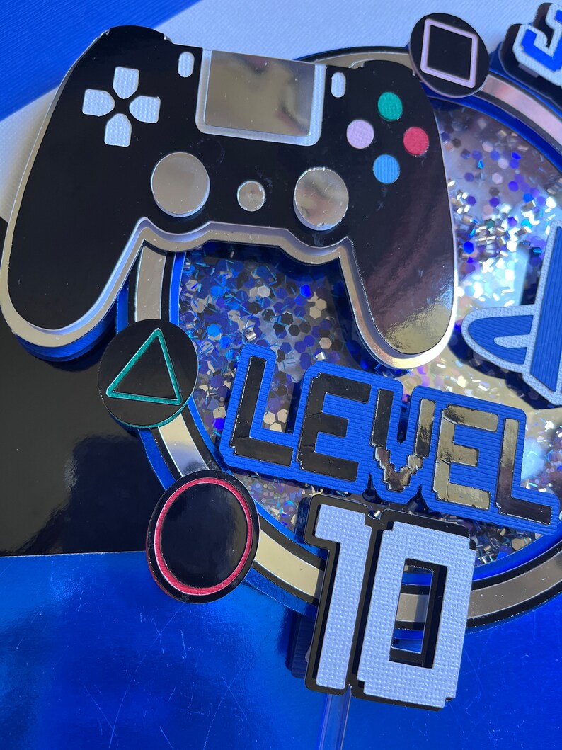 May include: A blue and silver cake topper with a black video game controller, a triangle, a circle, and the text "LEVEL 10".