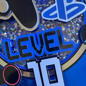 May include: A blue and black cake topper with a video game controller, a PlayStation logo, and the text "LEVEL 10".