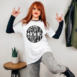 Könnte beinhalten: Ein weißes T-Shirt mit einem schwarzen Kreis-Motiv, auf dem in weißen Buchstaben "PEOPLE OVER PROFIT" steht. Die Person, die das Shirt trägt, ist eine Frau mit langen roten Haaren. Sie trägt eine schwarze Jeans und schwarze Stiefel.