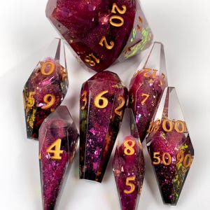 May include: A set of seven polyhedral dice with a coffin-like shape, crafted from clear resin containing deep magenta glitter and iridescent flakes. Each die features gold-colored numbers, including a large twenty-sided die at the top.