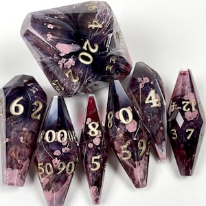 May include: A set of seven polyhedral dice featuring a translucent dark purple and black resin base with swirling pink and gold inclusions. Each die has sharp edges and is engraved with gold-colored numbers for clear readability.