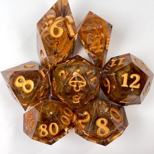 May include: A set of seven polyhedral dice made of translucent amber-colored resin with swirling gold glitter inclusions. Each die features engraved gold-colored numbers, and the twenty-sided die displays a small mushroom icon.
