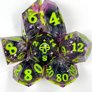 May include: A set of seven polyhedral dice made of translucent purple and black resin with swirling patterns, glitter, and bright neon green numbers. The twenty-sided die features a small mushroom icon in place of the number twenty.