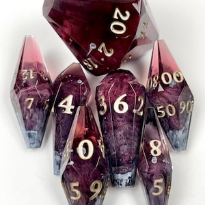 May include: A set of seven polyhedral dice with a deep burgundy and purple marbled resin finish. Each die features sharp edges and gold-colored engraved numbers. The collection includes a large twenty-sided die and six elongated, crystal-shaped dice with various numerical markings.