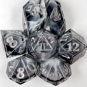 May include: A set of seven polyhedral dice made of clear resin with swirling black, white, and dark blue glitter inclusions. The numbers are embossed in silver, and the twenty-sided die features a small mushroom icon instead of the number twenty.