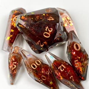 May include: A set of seven polyhedral dice made of clear resin with deep red and orange internal inclusions and metallic copper-colored numbers. The dice feature sharp edges and a glossy finish, with the twenty-sided die prominently displayed.
