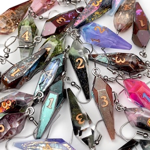 May include: A collection of multi-colored resin earrings shaped like elongated crystals or d4 dice. Each piece features a unique glitter or marbled finish, metallic silver-toned earring hooks, and a single engraved number painted in gold or silver.
