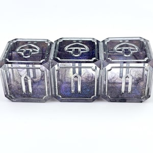 May include: Three clear, square boxes with silver trim and a decorative silver handle on top. The boxes have a dark, glittery interior with purple and silver hues. The boxes are arranged in a row against a white background.