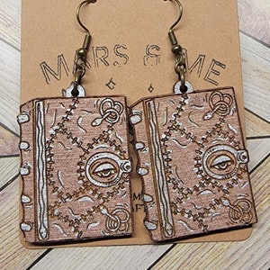 May include: A pair of wooden earrings shaped like a book with a white eye and gears design. The earrings are hanging from a brown card with the text "Mars & Me" printed on it.