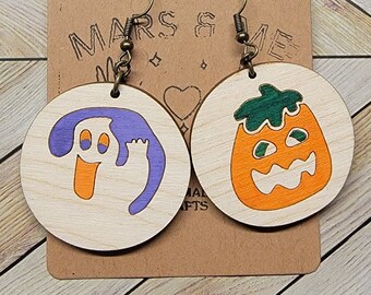 Könnte beinhalten: Ein Paar Halloween-Ohrringe mit einem Geist und einem Kürbis. Der Geist ist lila und weiß, der Kürbis orange und grün. Die Ohrringe sind aus Holz und haben ein schlichtes Design.