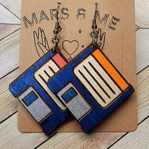 Peut inclure: Une paire de boucles d'oreilles en forme de disquette bleue et orange avec des accents argentés. Les boucles d'oreilles sont sur une carte brune avec le texte "MARS & ME" imprimé dessus.