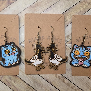 May include: Three pairs of novelty earrings displayed on brown card backings. The earrings feature cartoon cat and bird designs in blue, brown, black, and white. The cat earrings have a blue body, and the birds wear hats. The earrings hang from bronze-coloured hooks.