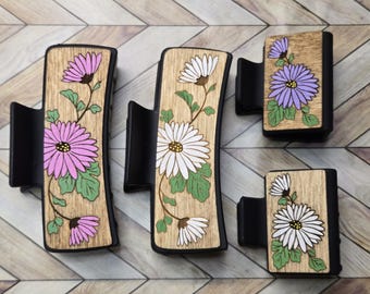 May include: Three wooden hair clips with floral designs. The clips are black and have a curved shape. The floral designs are of white, pink, and purple flowers with green leaves.
