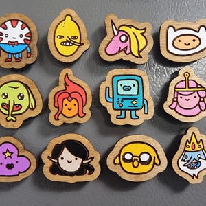 Könnte beinhalten: Eine Sammlung von Adventure Time Holzmagneten mit verschiedenen Charakteren. Jeder Magnet ist einzigartig geformt und handbemalt mit leuchtenden Farben, darunter Finn, Jake und Prinzessin Bubblegum. Die Magnete sind aus Holz mit natürlicher Maserung.