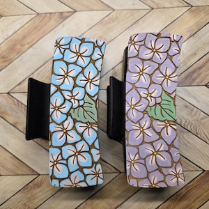 May include: Two rectangular hair clips with black bases. One clip features a light blue floral design, and the other has a purple floral pattern. Both have brown outlines and green leaves.