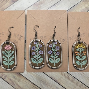 May include: Three pairs of floral earrings displayed on brown cards. Each earring features a brown oval with a silver frame and a colorful floral design. The flowers are pink, purple, and yellow with green leaves.