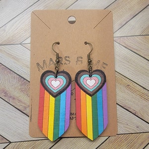 May include: A pair of wooden earrings featuring a rainbow design. Each earring has a heart-shaped top with a heart-within-a-heart design, and a rainbow-colored fringe. The earrings are attached to bronze-colored ear wires. The earrings are displayed on a brown card with the text "MARS & ME".