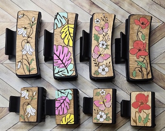 May include: Eight wooden hair clips with different floral and leaf designs. The clips are black with a wood insert featuring a variety of flowers, butterflies, and leaves. The designs include white bells, pink and blue tropical leaves, white flowers with pink butterflies, and red poppies.