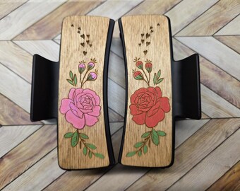May include: Two wooden hair clips with a floral design. The clips are black and have a wooden insert with a pink and red rose design. The roses are surrounded by small green leaves and stems.